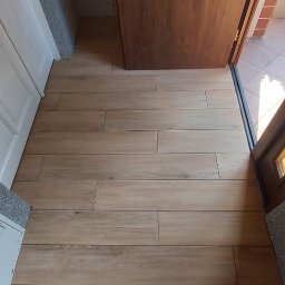 Budowa domów pasywnych Barbara i Bogdan Łabęccy spółka cywilna - Ułożona podłoga z płytek imitujących drewno w jasnym odcieniu, widok z perspektywy osoby stojącej w szarych kapciach, z otwartymi drzwiami wejściowymi.