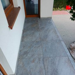 Budowa domów pasywnych Barbara i Bogdan Łabęccy spółka cywilna - Wyłożona płytkami podłoga balkonu z widoczną fakturą kamienia, przy białej ścianie budynku z drewnianą ramą okienną. Na zewnątrz kostka brukowa i zieleń.