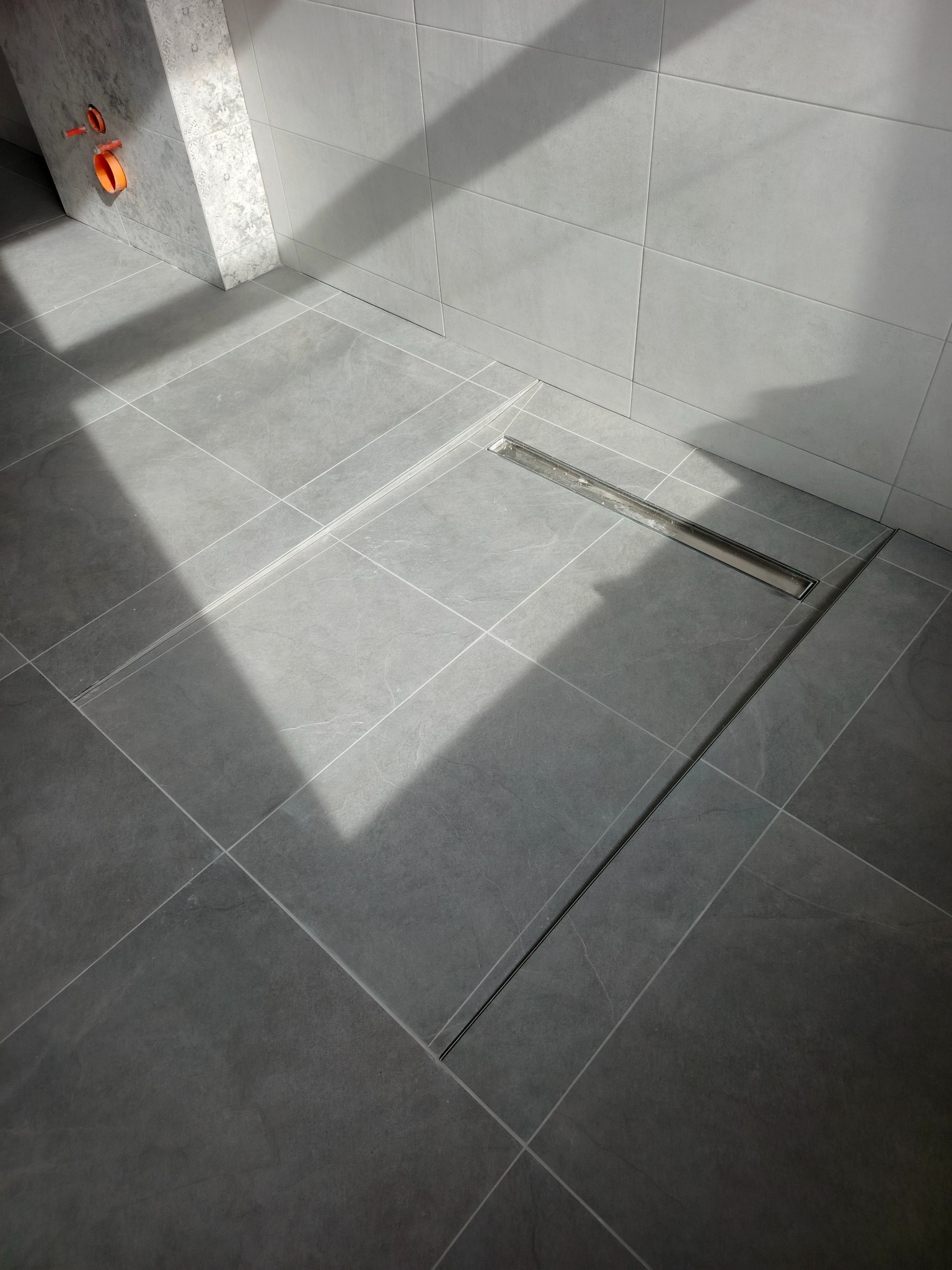 Nowoczesna łazienka z szarymi płytkami i odpływem liniowym. Widoczne fragmenty instalacji. Minimalistyczny design, jasne wnętrze z geometrycznym wzorem płytek.