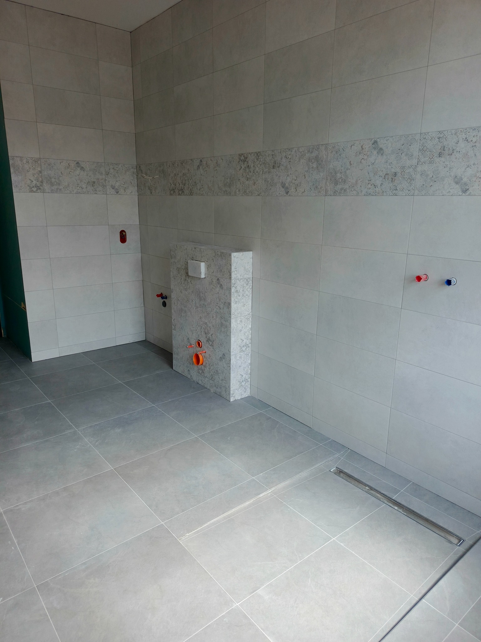 Wykończona łazienka w szarościach: płytki na ścianach z dekoracyjnym pasem, podłoga wyłożona gresem, widoczny odpływ liniowy i zabudowa stelaża WC z przyłączami.