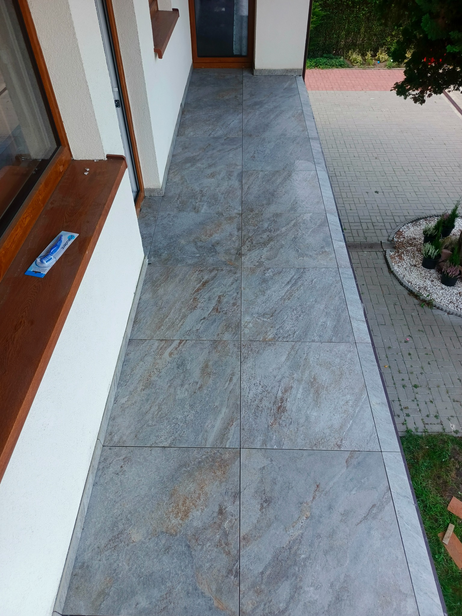 Wykończony balkon z szarymi płytkami imitującymi kamień. Widok z góry ukazuje estetyczne fugi i detale wykończenia, z zewnątrz widoczny fragment chodnika z kostki.