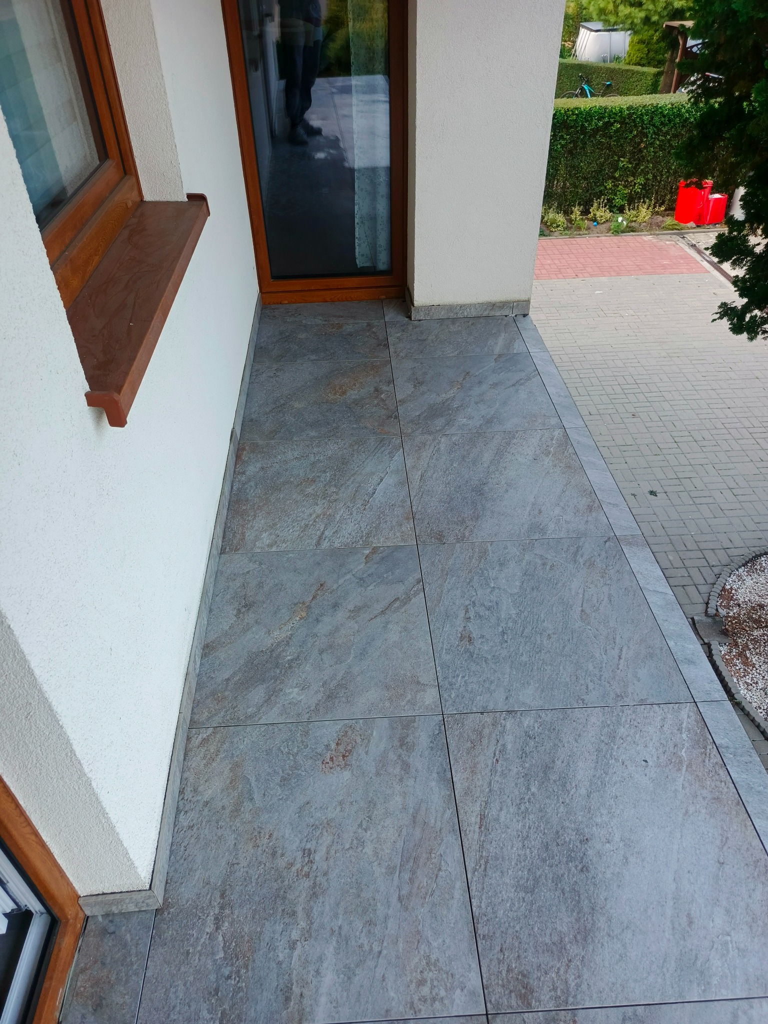 Wyłożona płytkami podłoga balkonu z widoczną fakturą kamienia, przy białej ścianie budynku z drewnianą ramą okienną. Na zewnątrz kostka brukowa i zieleń.