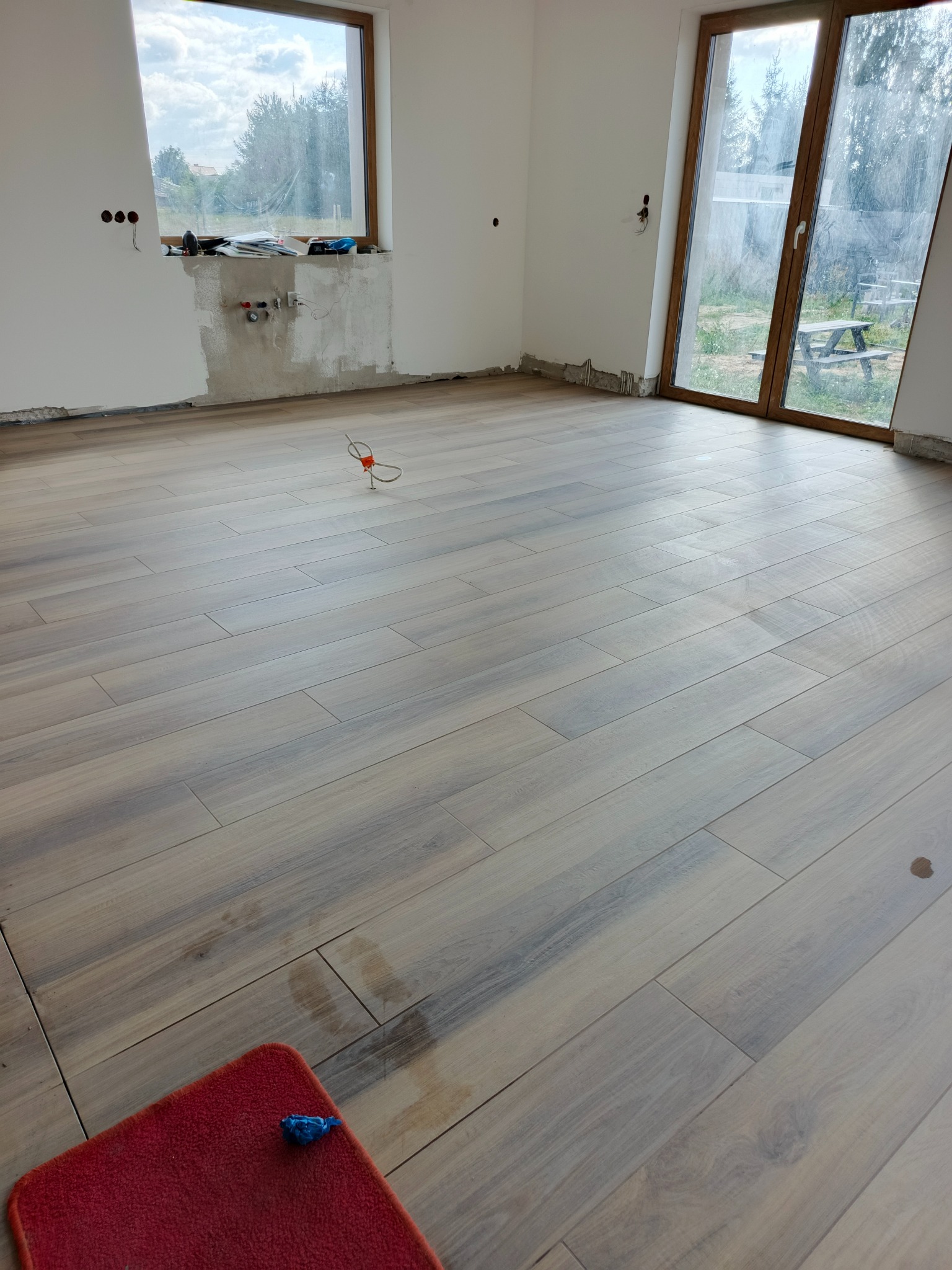 Ułożona podłoga z płytek imitujących drewno w jasnym wnętrzu. Widoczne ślady po budowie, czerwony dywanik i wystające kable. Duże okno z widokiem na zewnątrz.