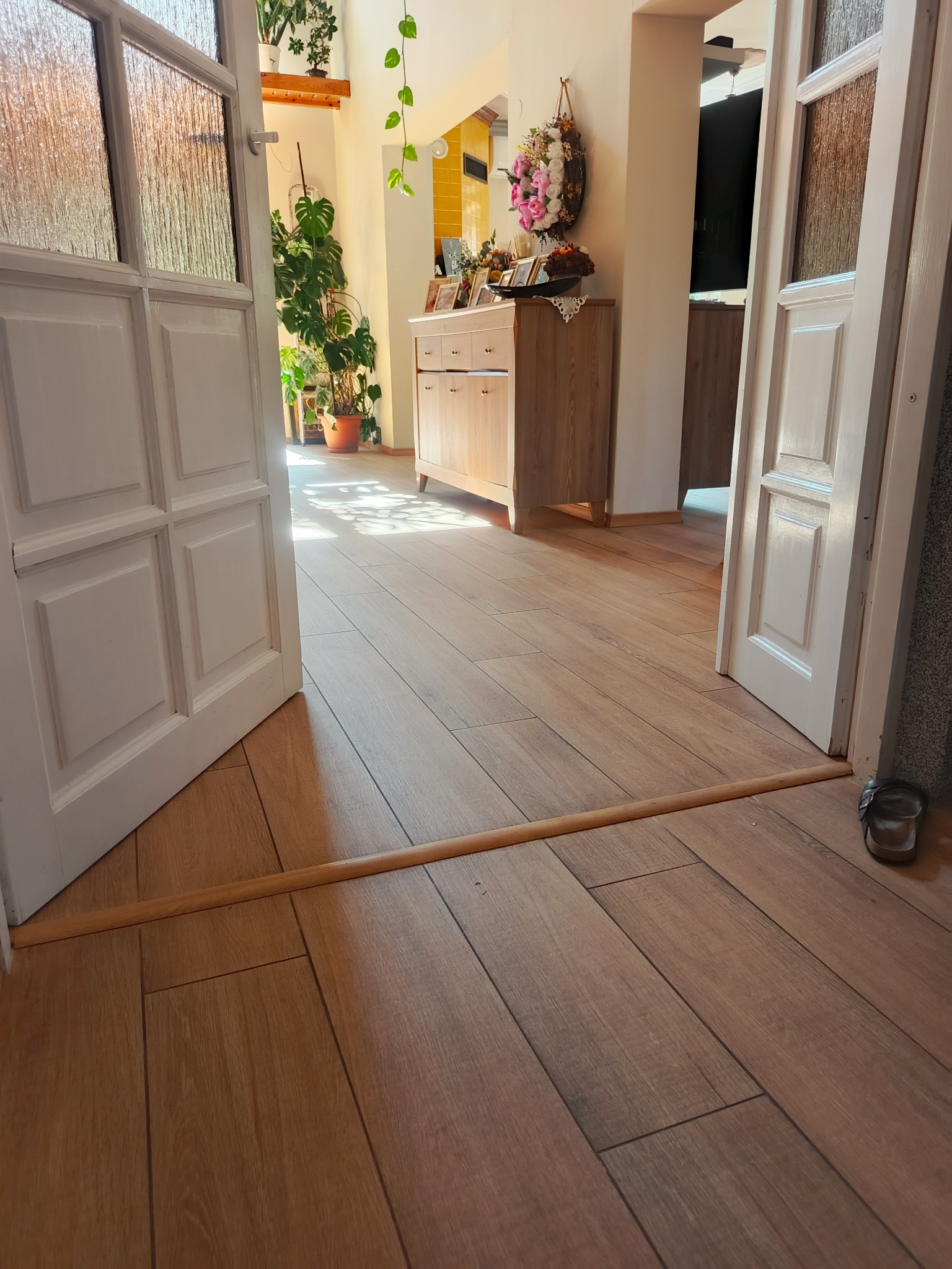 Jasne, drewnopodobne płytki podłogowe ułożone w korytarzu, widoczne przez otwarte, białe drzwi. W tle komoda i dekoracje roślinne.