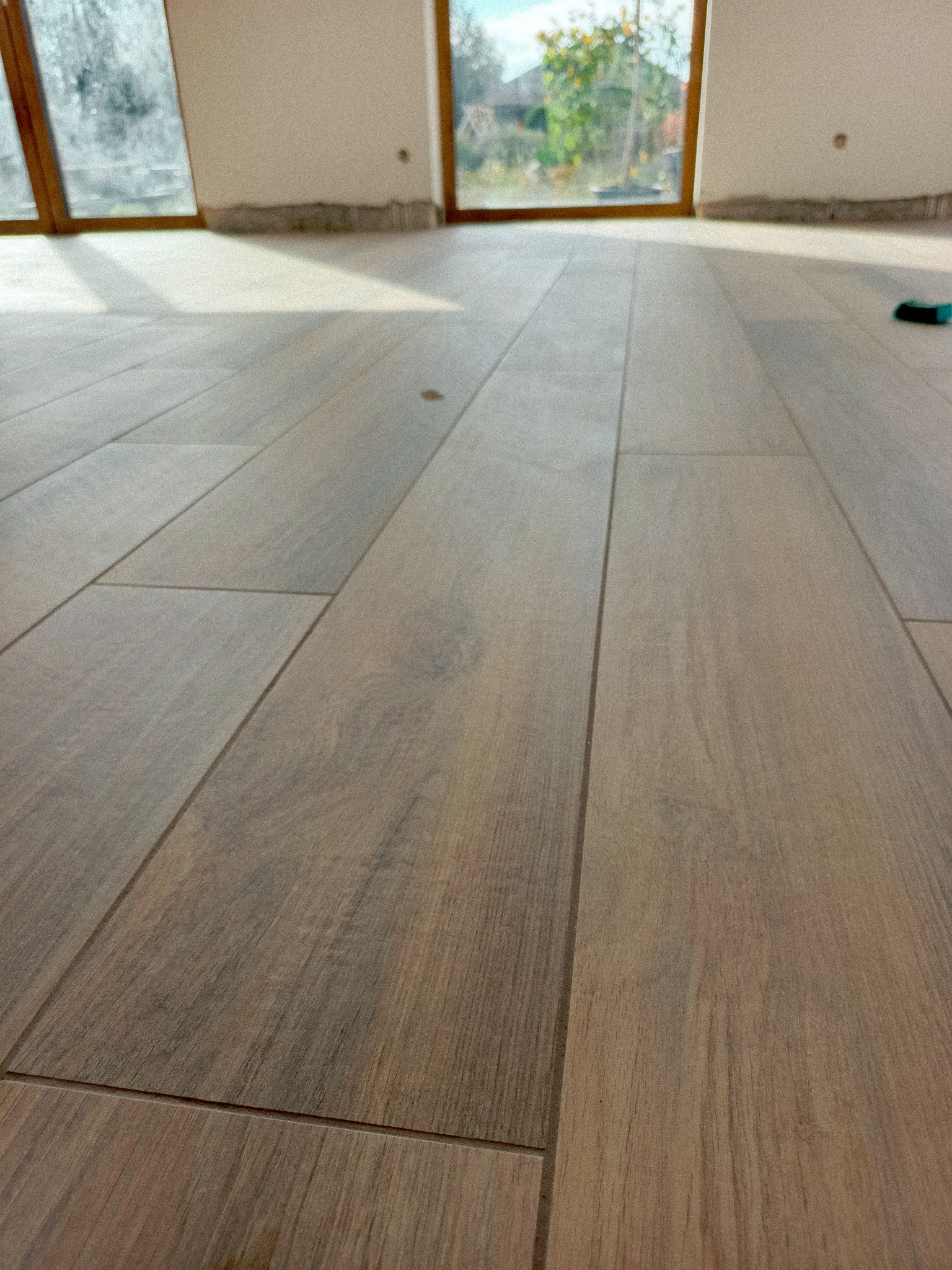 Ułożone, podłużne płytki podłogowe imitujące drewno, z widocznym oknem w tle i narzędziem glazurniczym. Jasne wnętrze, naturalne światło.