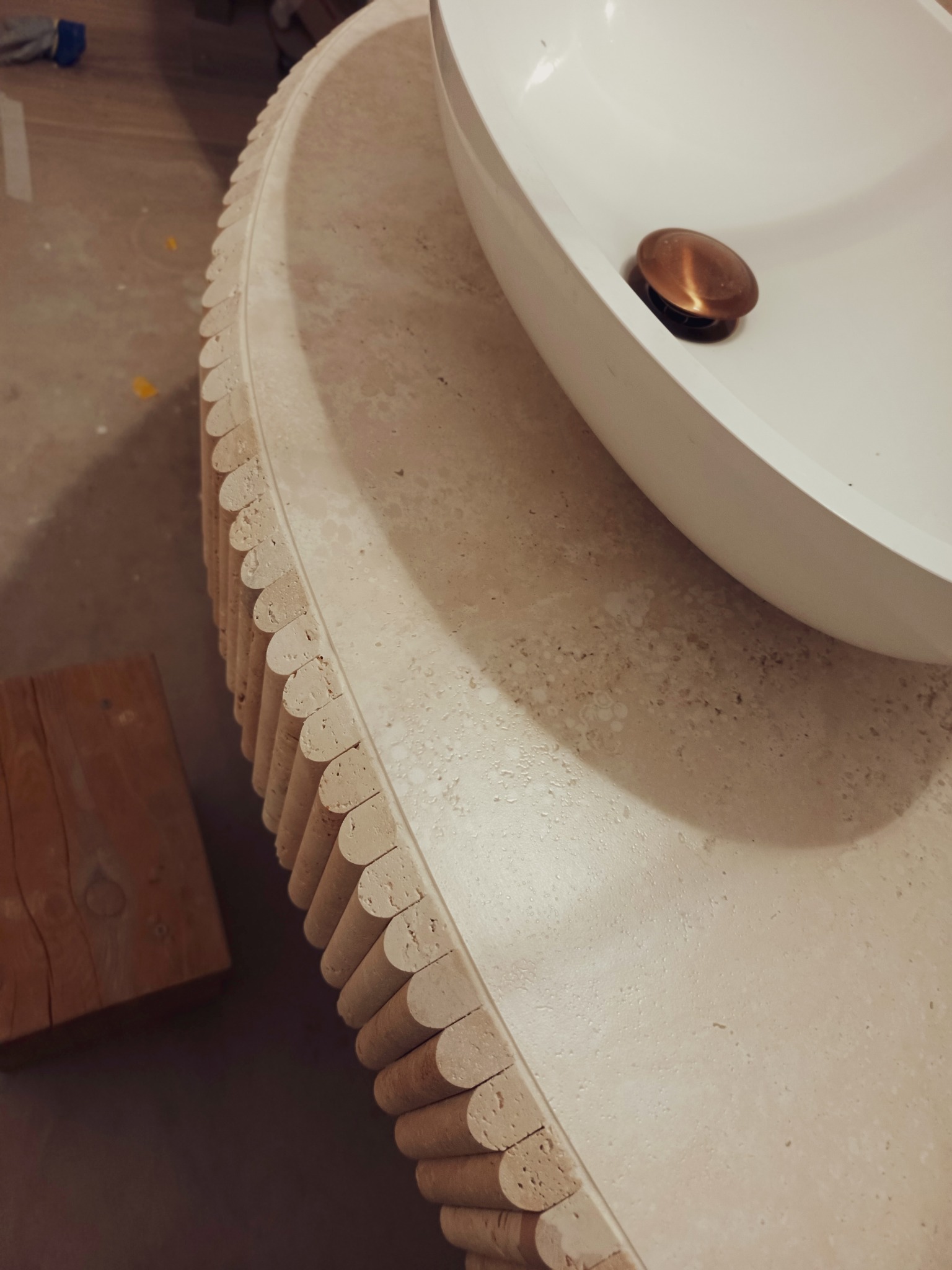 Owalna umywalka nablatowa z mosiężnym korkiem, osadzona na blacie z trawertynu z dekoracyjnym frezem. Widoczny fragment drewnianego klocka.