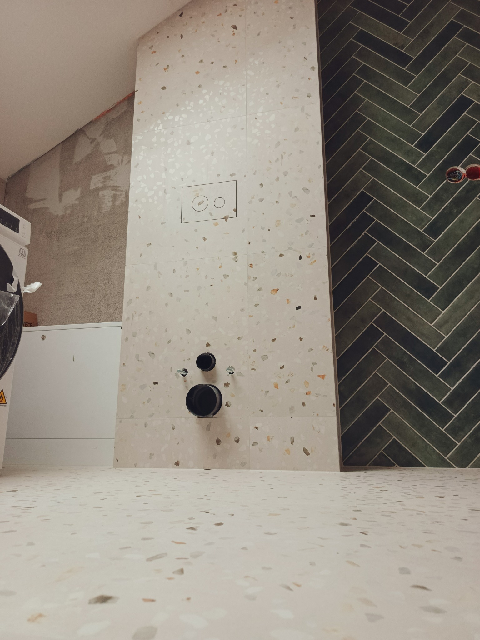 Ściana łazienki wyłożona płytkami w stylu lastryko i ciemnozieloną jodełką. Widoczne elementy instalacji sanitarnej i fragment pralki. Ukończony etap prac.