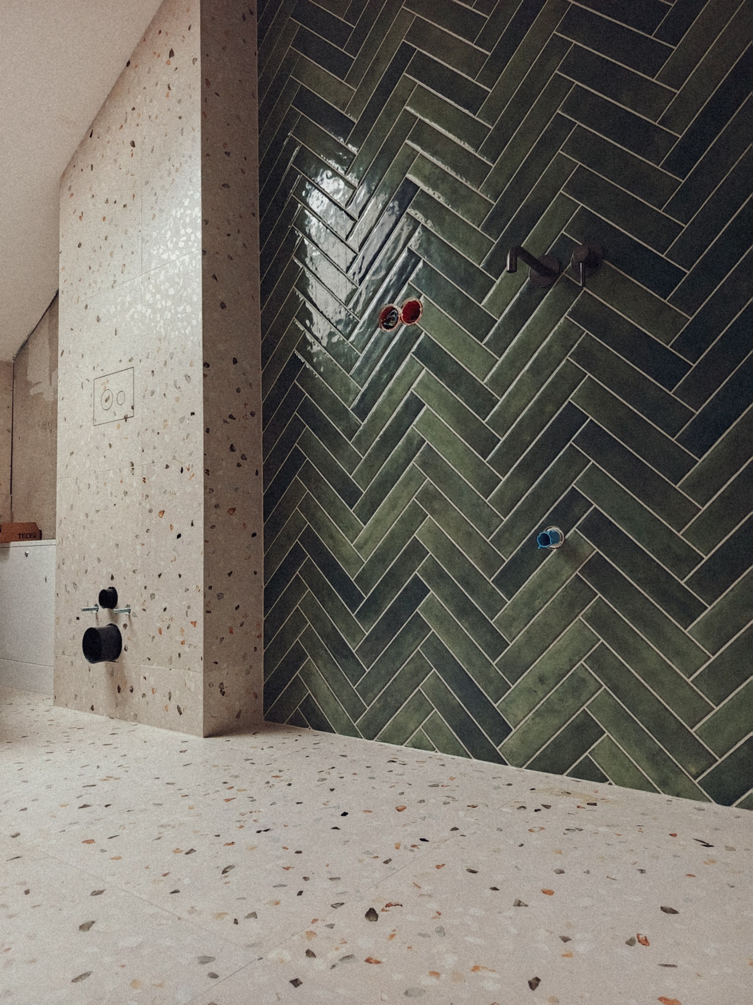 Nowoczesna łazienka z zielonymi płytkami ułożonymi w jodełkę na ścianie i podłogą wyłożoną płytkami lastryko w odcieniach beżu i brązu. Widoczne elementy instalacji.