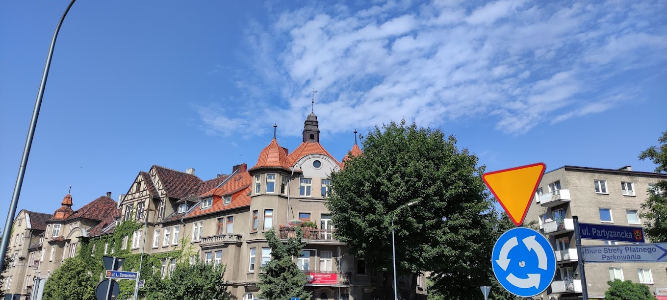 Odnowiona kamienica z wieżyczką i czerwoną dachówką w Raczycach, z elementami muru pruskiego, porośnięta bluszczem, na tle błękitnego nieba z chmurami.