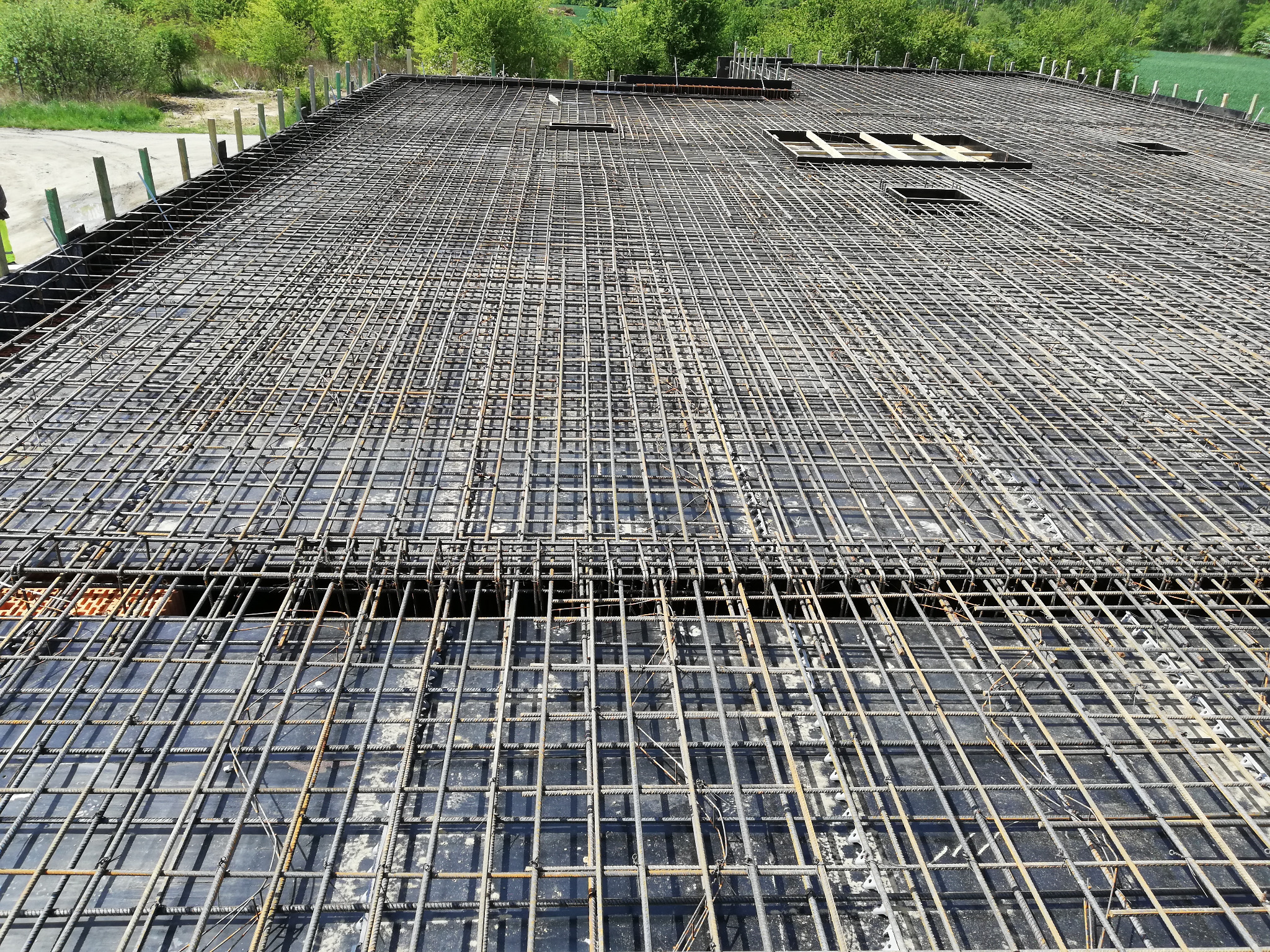 Siatka zbrojeniowa na czarnym podłożu, przygotowana do zalania betonem, widok z góry na budowie budynku żelbetowego, w tle zieleń drzew.