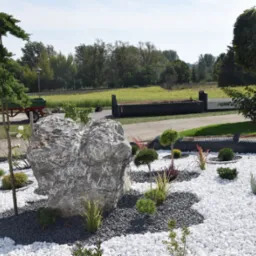 Ogród ozdobny z dużym szarym kamieniem, białymi i szarymi kamykami, niskimi krzewami i przyciętymi drzewkami, na tle widoczna przyczepa z zieloną skrzynią i drzewa