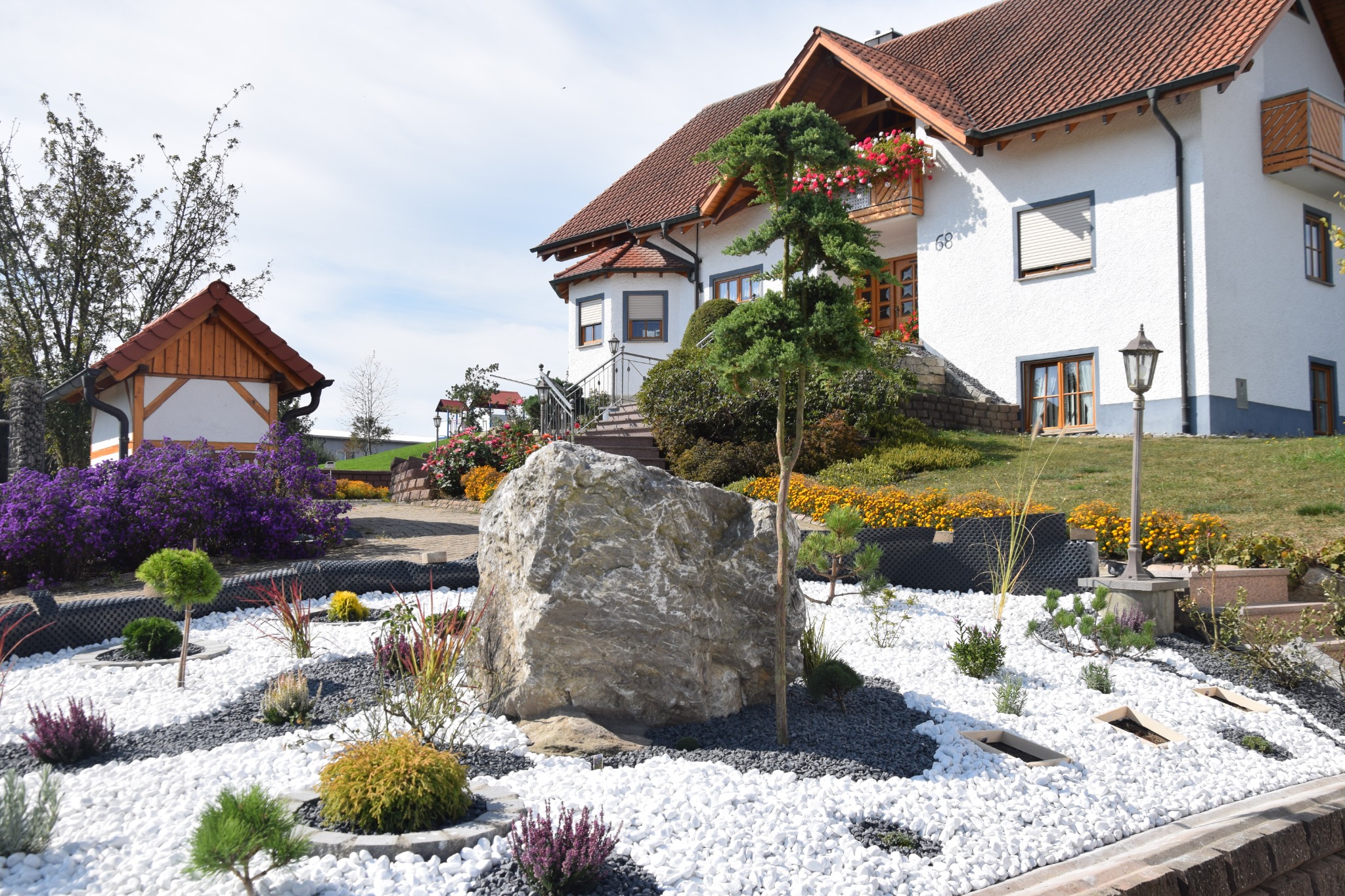 Ogród przydomowy z białymi i szarymi kamieniami, ozdobnymi krzewami i drzewkami iglastymi, na tle białego domu z czerwoną dachówką i kwiatami na balkonie.