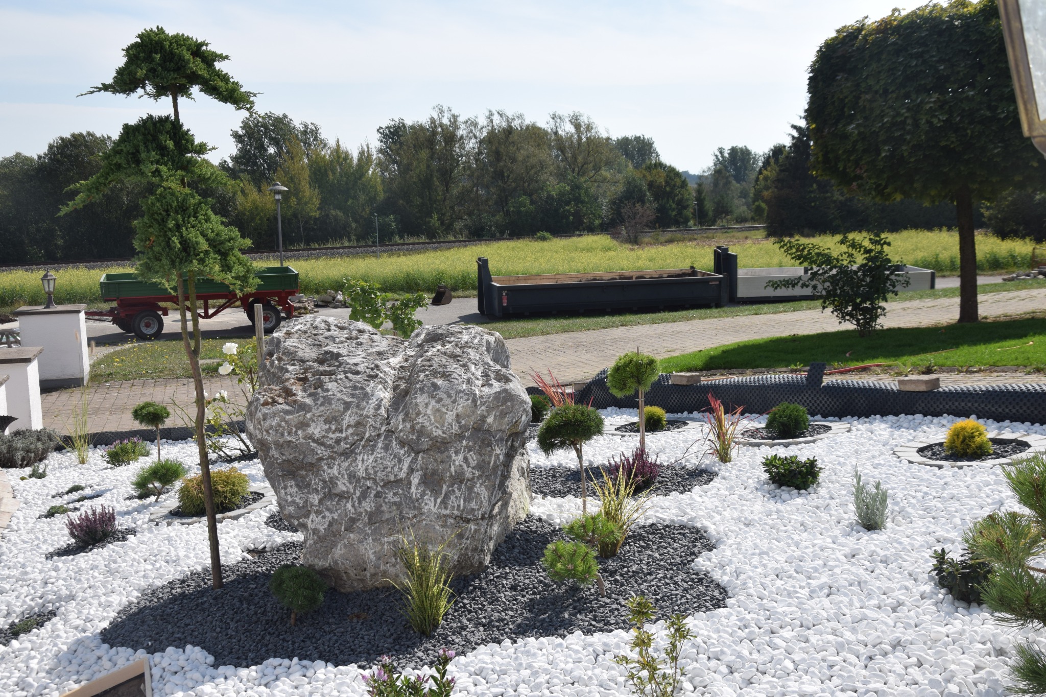 Ogród ozdobny z dużym szarym kamieniem, białymi i szarymi kamykami, niskimi krzewami i przyciętymi drzewkami, na tle widoczna przyczepa z zieloną skrzynią i drzewa