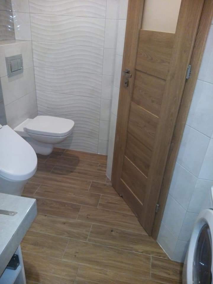 Nowoczesna łazienka z toaletą podwieszaną, płytkami imitującymi drewno na podłodze i ścianie, oraz drewnianymi drzwiami.