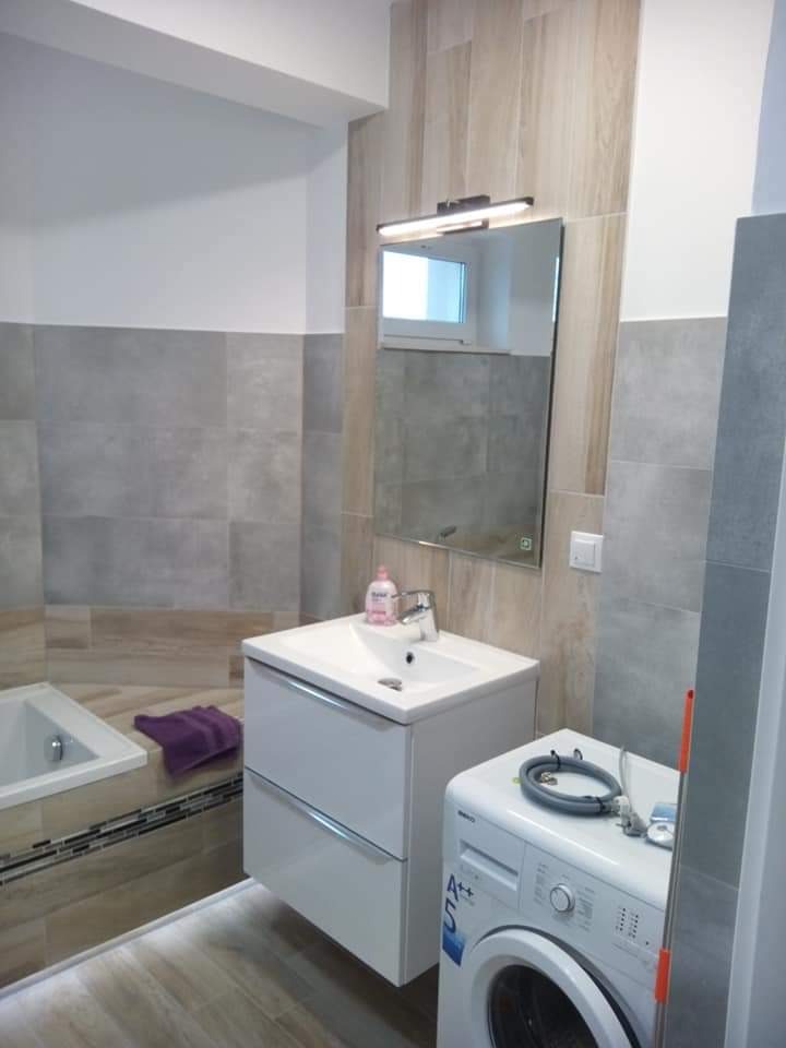 Nowoczesna łazienka po remoncie: biała umywalka z szafką, lustro z oświetleniem, pralka z widocznym wężem, wanna z mozaiką i fioletowy ręcznik, płytki imitujące drewno na ścianach i podłodze.