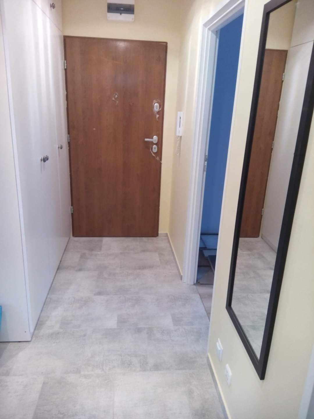 Wykończony korytarz z szarymi płytkami podłogowymi, białą szafą w zabudowie, drewnianymi drzwiami wejściowymi i lustrem w czarnej ramie.