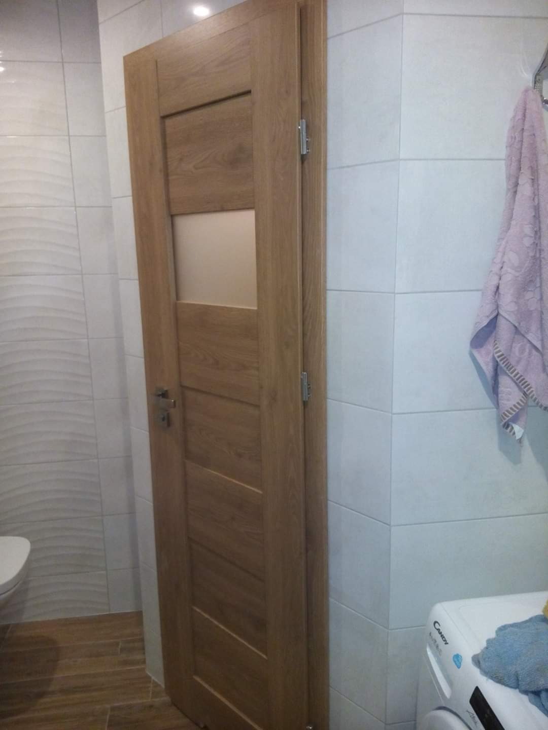 Zamknięte drewniane drzwi z przeszkleniem w łazience z białą, wzorzystą glazurą i fragmentem pralki oraz ręcznikiem.