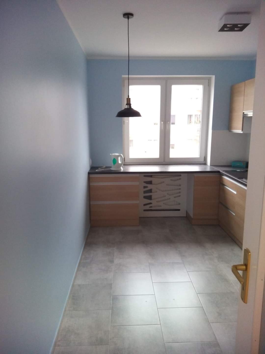 Jasna kuchnia z szarymi płytkami podłogowymi, drewnianymi szafkami, czarnym blatem i oknem z widokiem na zewnątrz, oświetlona wiszącą lampą w stylu industrialnym.