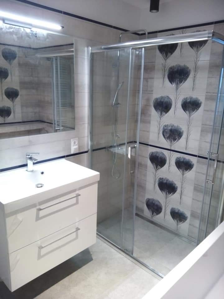 Nowoczesna łazienka z kabiną prysznicową ze szklanymi drzwiami przesuwnymi, białą szafką pod umywalkę z dwiema szufladami i płytkami z motywem kwiatowym na ścianie.