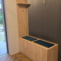 SEBEX Sebastian Chojnacki - Funkcjonalna szafka z siedziskiem i wieszakami w przedpokoju. Drewnopodobny dekor, pionowe panele na ścianie, podłoga ułożona w jodełkę. Widok na zewnątrz przez otwarte drzwi.