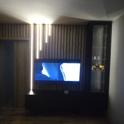 SEBEX Sebastian Chojnacki - Nowoczesny salon z lamelami na ścianie, podświetleniem LED i szafką RTV. W szafce szklanej widoczny model traktora. Minimalistyczny design.