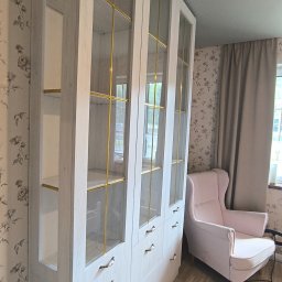 SEBEX Sebastian Chojnacki - Biała szafa na wymiar z przeszklonymi witrynami i złotymi akcentami, dopasowana do wnętrza z tapetą w kwiaty i jasną podłogą, obok różowy fotel.