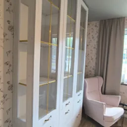 Biała szafa na wymiar z przeszklonymi witrynami i złotymi akcentami, dopasowana do wnętrza z tapetą w kwiaty i jasną podłogą, obok różowy fotel.