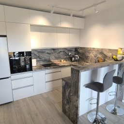 SEBEX Sebastian Chojnacki - Nowoczesna kuchnia na wymiar z białymi frontami, blatem imitującym marmur i barkiem z dwoma hokerami. Oświetlenie punktowe.