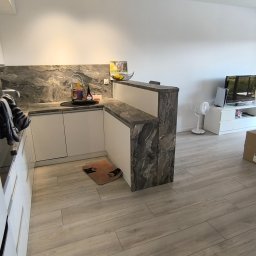 SEBEX Sebastian Chojnacki - Nowoczesna kuchnia z białymi szafkami i blatem imitującym marmur, widoczna z perspektywy wejścia do pokoju. Uwagę przykuwa wykończenie półwyspu marmurkowym wzorem.