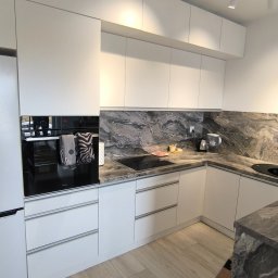 SEBEX Sebastian Chojnacki - Nowoczesna, biała kuchnia z blatem imitującym marmur, oświetleniem punktowym i drewnianą podłogą. Widoczny piekarnik z ręcznikami i elementy wyposażenia.