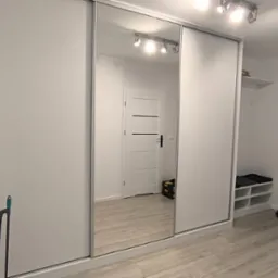 Biała szafa wnękowa z lustrzanymi drzwiami przesuwnymi, odbijająca wnętrze przedpokoju z białą podłogą i drzwiami. Minimalistyczny design.