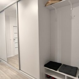 SEBEX Sebastian Chojnacki - Biała szafa wnękowa z lustrzanymi drzwiami przesuwnymi w przedpokoju. Obok wieszak z siedziskiem i półkami na buty. Minimalistyczny design, jasne wnętrze.