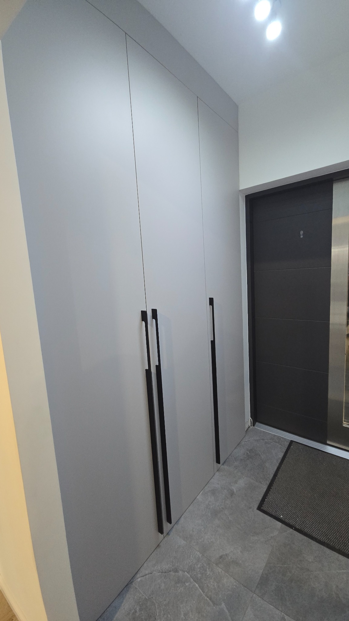 Jasnoszara szafa wnękowa z długimi, pionowymi, czarnymi uchwytami w przedpokoju z szarą podłogą i ciemnymi drzwiami wejściowymi.