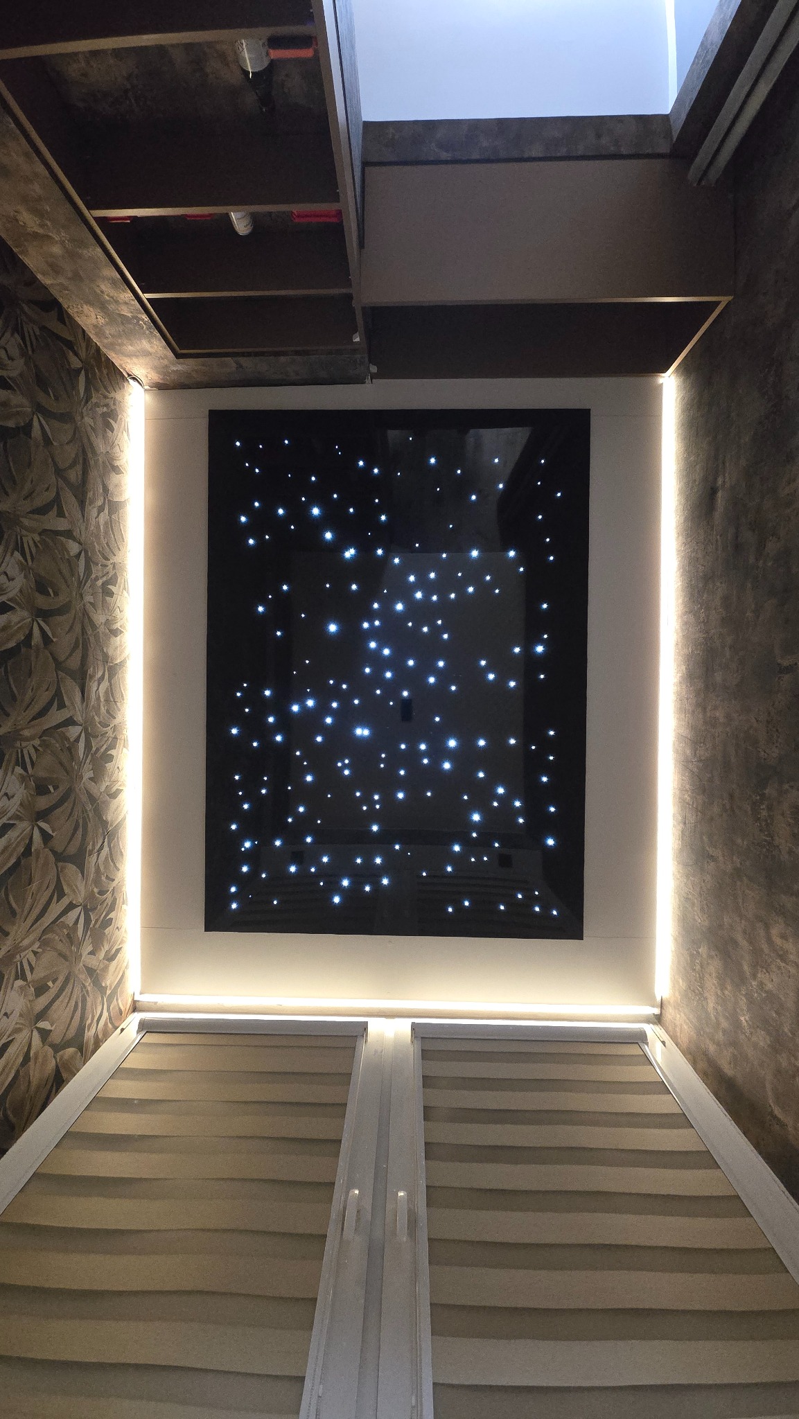 Nowoczesny sufit z efektem gwieździstego nieba, podświetlony LED, z żaluzjami w dolnej części kadru i dekoracyjną tapetą po lewej stronie. Unikalny design wnętrza.