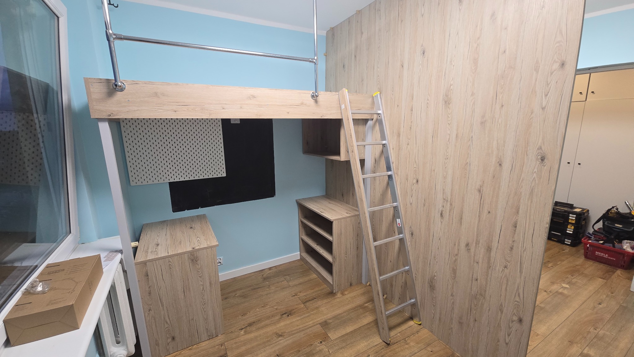 Funkcjonalna antresola z drabinką i szafkami w pokoju o błękitnych ścianach. Jasne drewno, minimalistyczny design, widoczne narzędzia w tle.