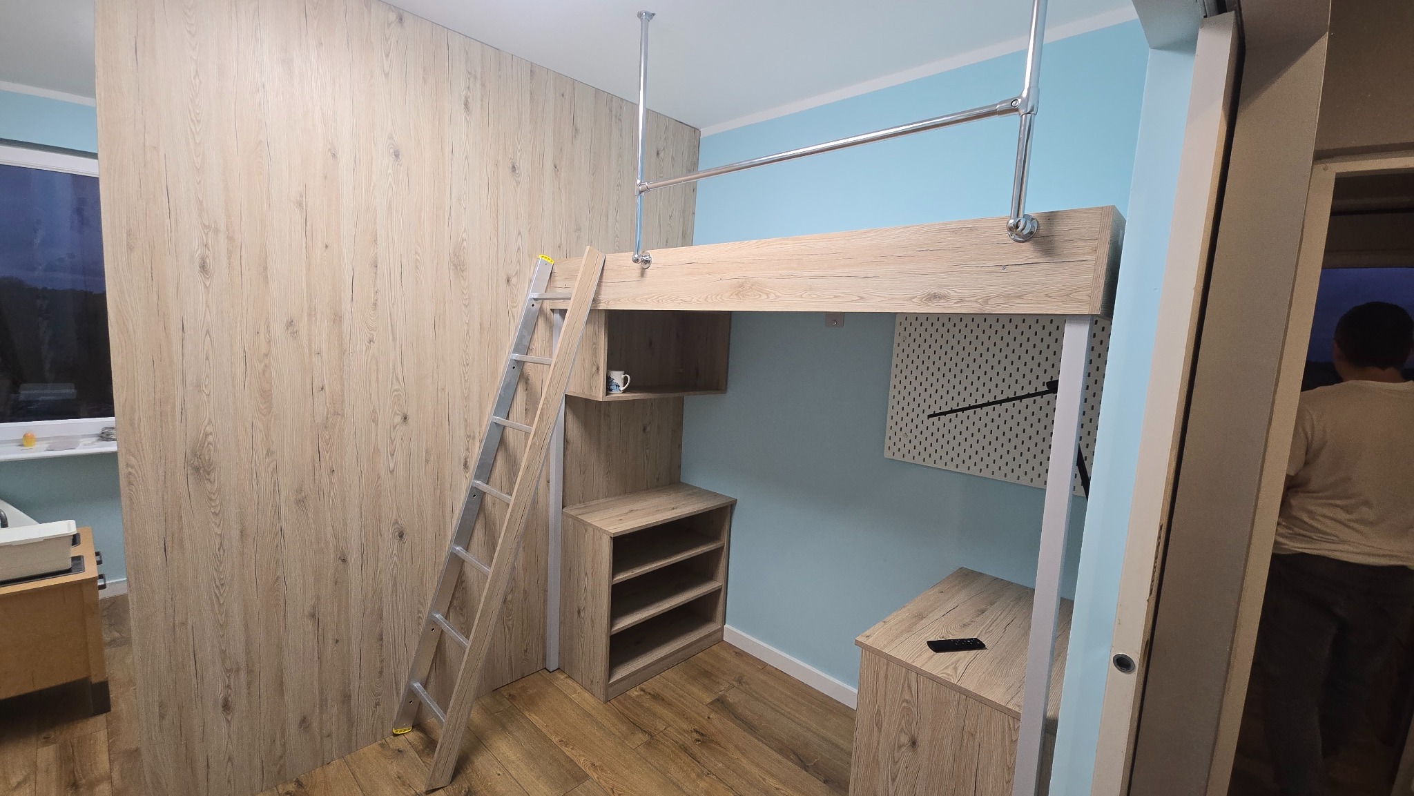 Drewniana antresola z drabinką i szafkami w pokoju o błękitnych ścianach. Metalowy drążek do wieszania ubrań. Widoczna część osoby w tle.