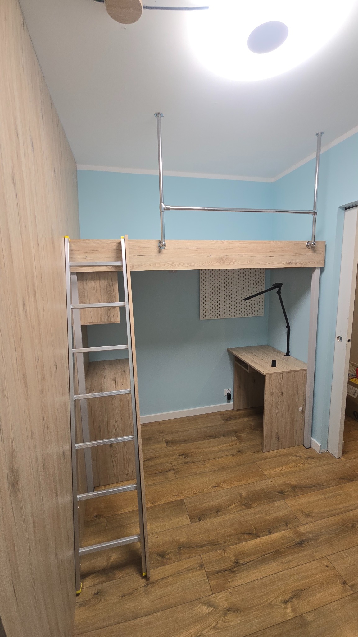 Funkcjonalna antresola z biurkiem i drabinką w pokoju dziecięcym. Jasne drewno, błękitne ściany, metalowe rury podtrzymujące konstrukcję. Nowoczesny design, oszczędność miejsca.