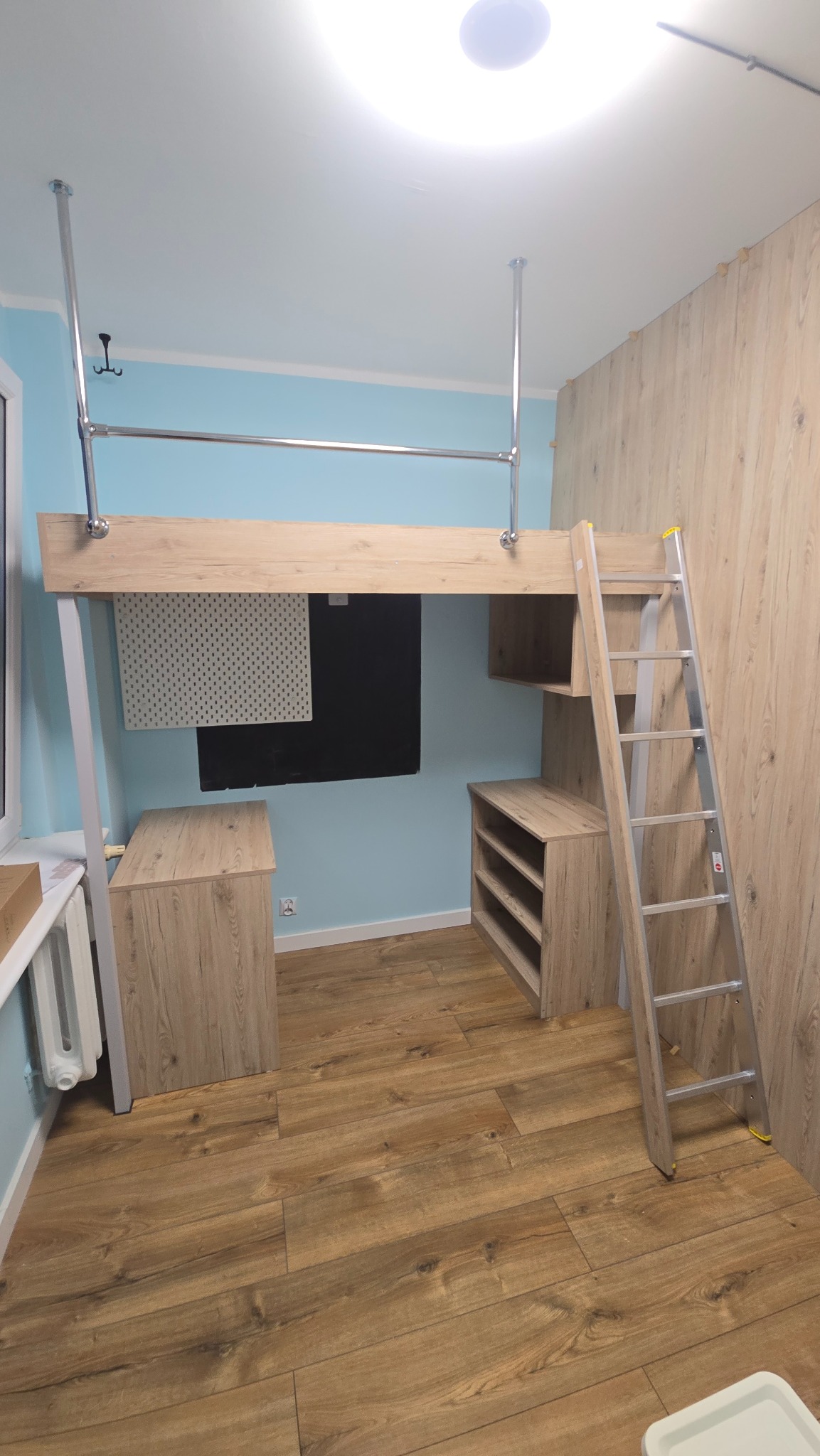 Pokój z antresolą w jasnym drewnie, metalowa drabinka, biurko i szafka w tym samym stylu. Niebieskie ściany, drewniana podłoga i częściowo drewniana ściana.