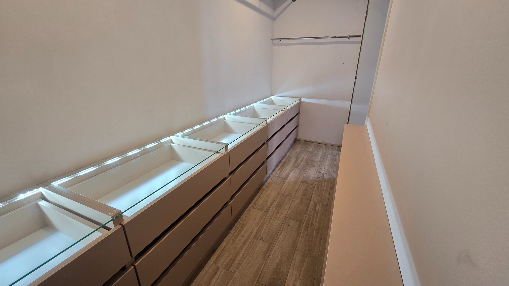 Beżowa szafa wnękowa z podświetleniem LED i szklanymi półkami w trakcie montażu, widok z perspektywy, drewniana podłoga, metalowy drążek na ubrania.