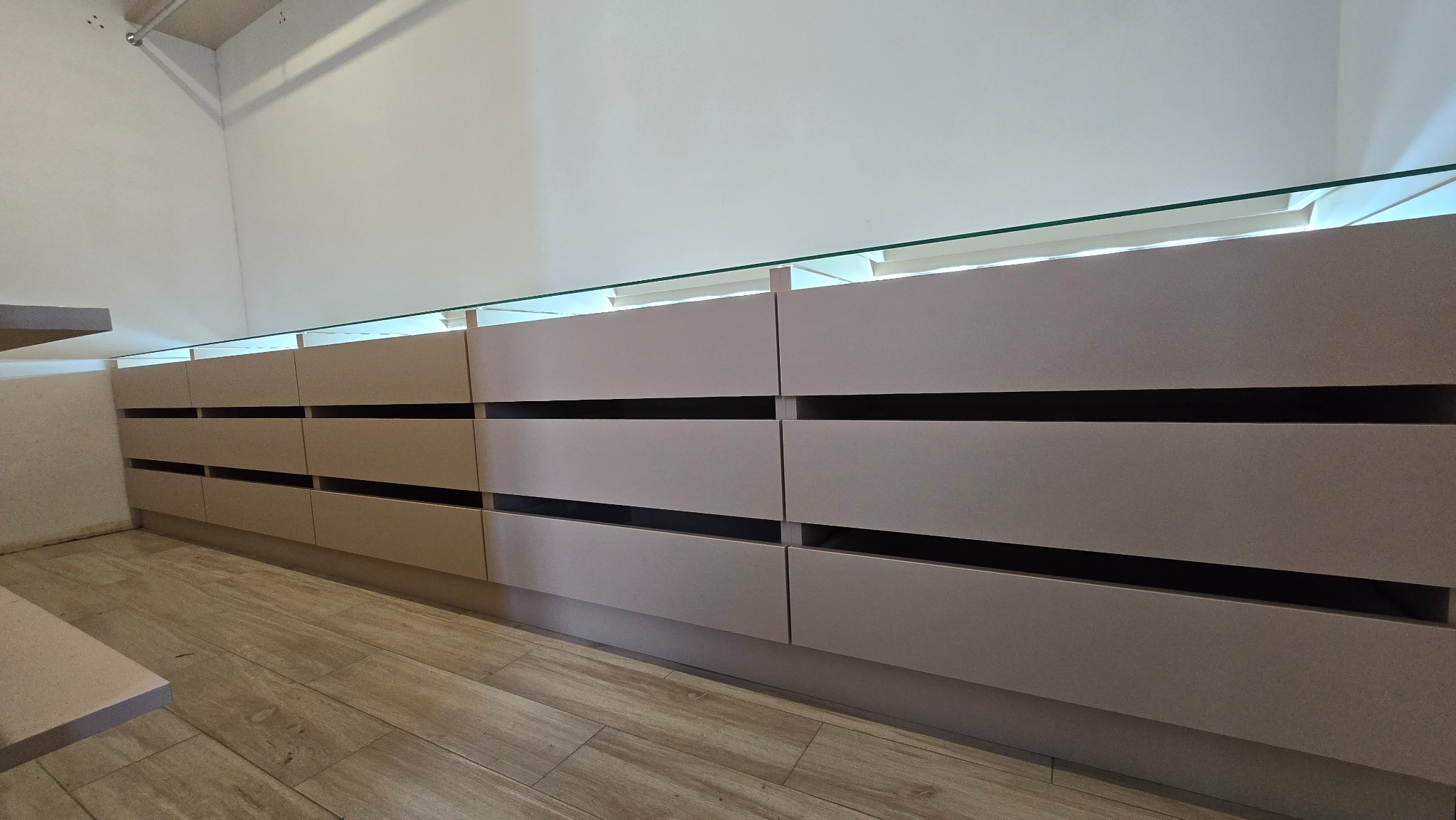 Beżowa szafa wnękowa z podświetleniem LED i przeszklonym blatem, widoczna z perspektywy podłogi, na tle białej ściany i drewnianej podłogi.