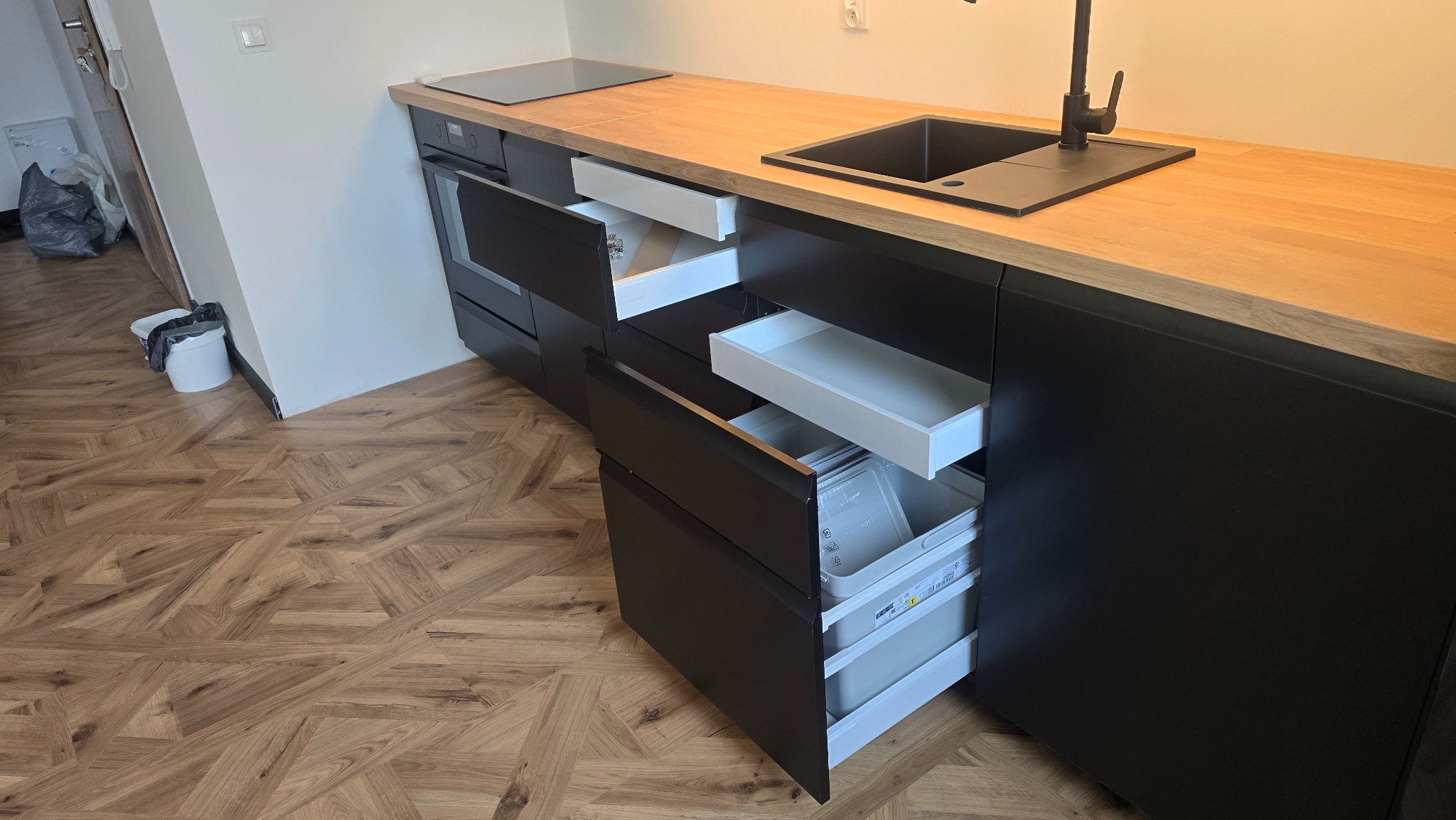 Nowoczesna kuchnia z czarnymi frontami i drewnianym blatem. Otwarta szuflada z pojemnikami. Widoczny zlew z czarną armaturą i płyta grzewcza.