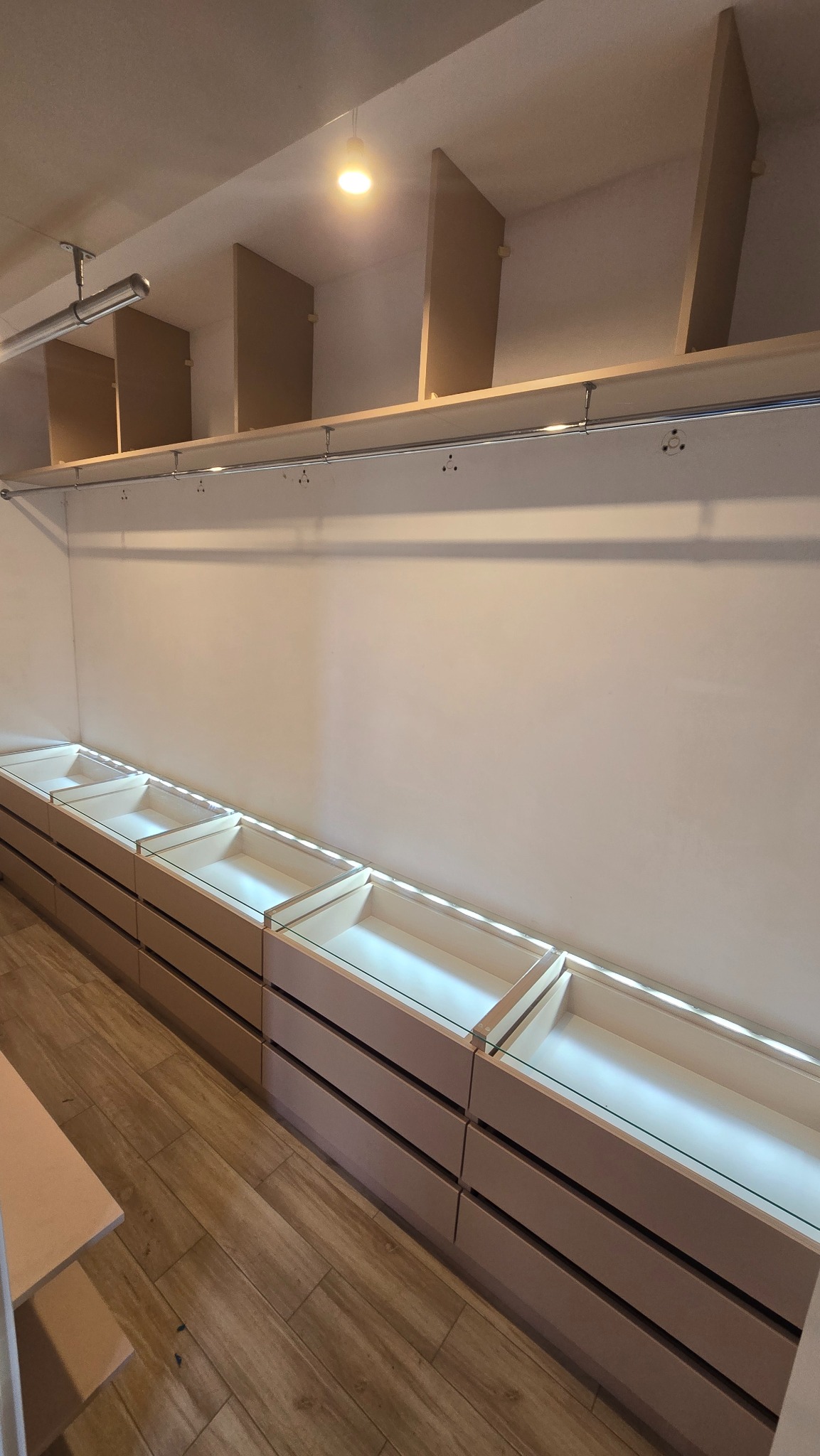 Beżowa szafa wnękowa z szufladami, oświetleniem LED i półkami pod sufitem. Widok z dołu, ukazujący detale wykończenia i przestrzeń do przechowywania.