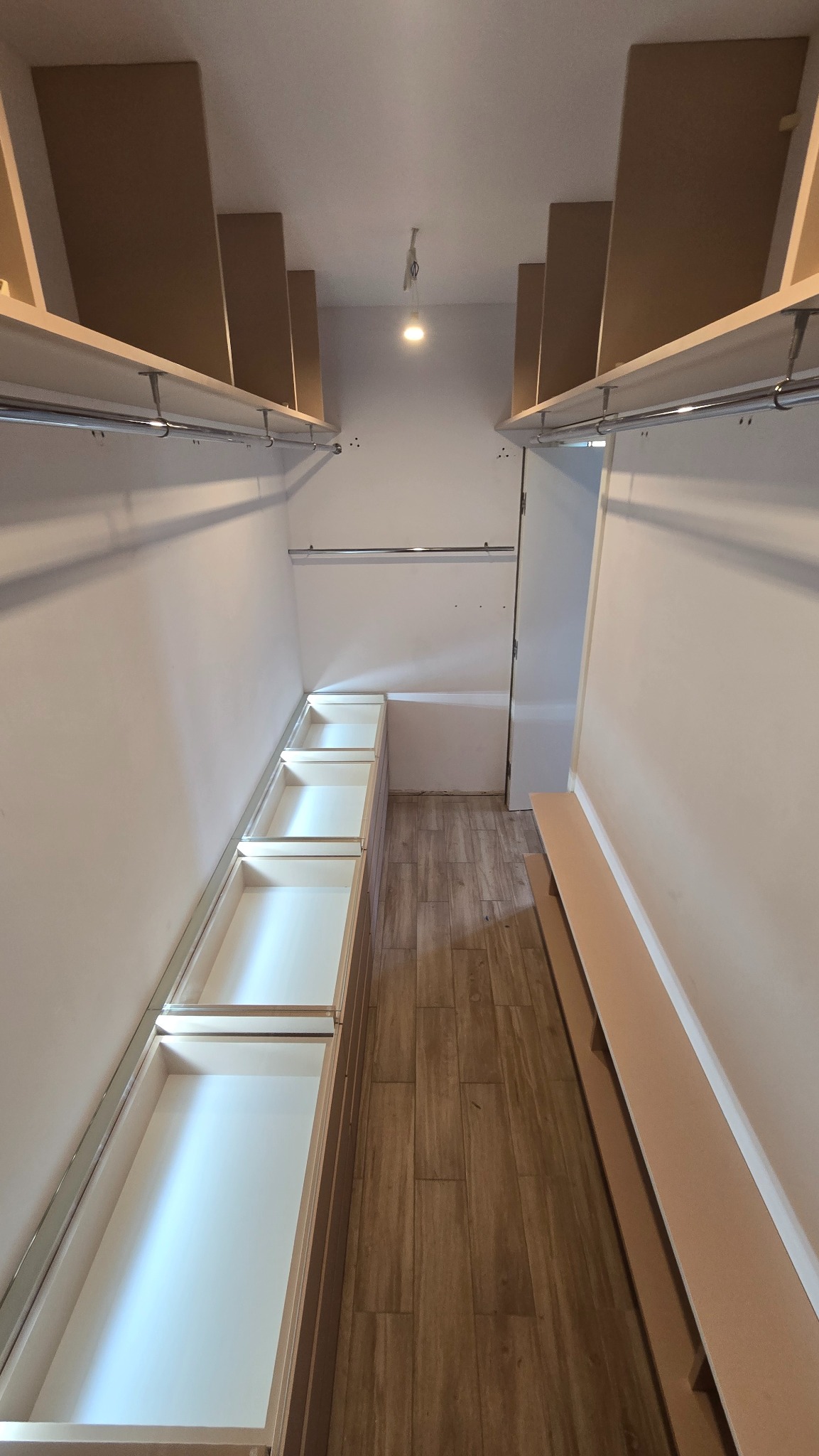 Wnętrze garderoby z szufladami i półkami, widok z dołu. Białe fronty szuflad kontrastują z drewnianą podłogą. Metalowe drążki na ubrania. Oświetlenie sufitowe.
