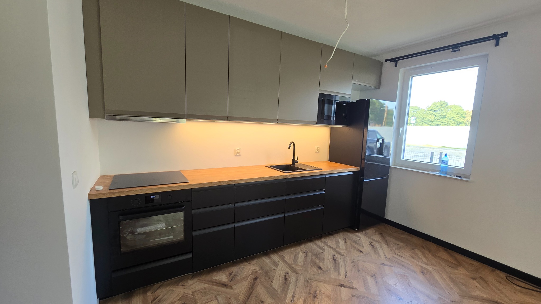 Nowoczesna kuchnia z szarymi szafkami górnymi i czarnymi dolnymi, drewnianym blatem, czarną armaturą i wzorzystą podłogą. Widok na okno z widokiem na zieleń.