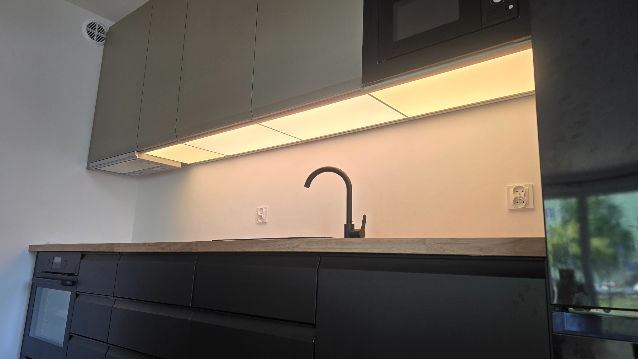 Nowoczesna, czarna kuchnia z drewnianym blatem i podświetleniem LED pod szafkami. Widoczny zlew, bateria i sprzęt AGD w zabudowie. Minimalistyczny design.