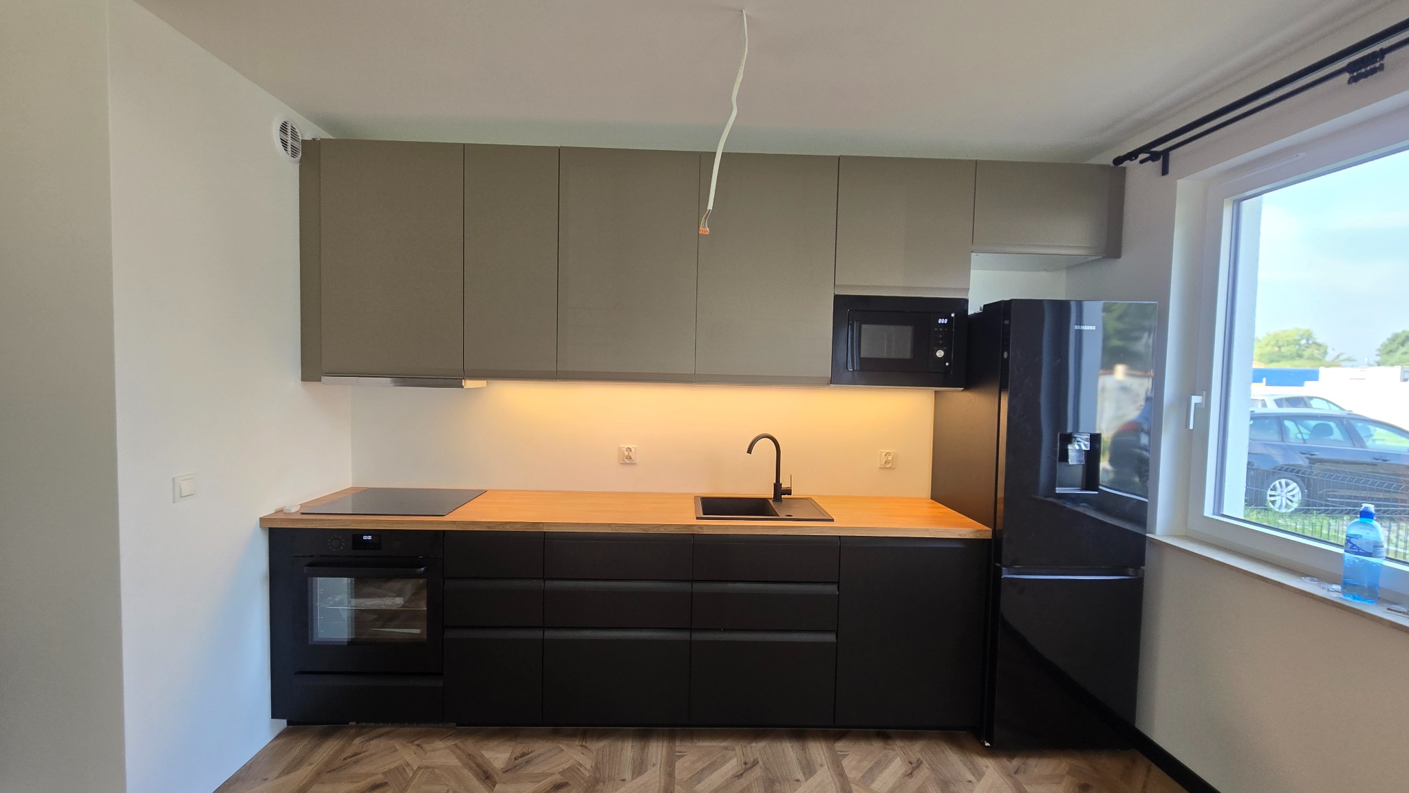 Nowoczesna kuchnia z czarnymi szafkami dolnymi, drewnianym blatem i szarymi szafkami górnymi. Widoczna lodówka i okno z widokiem na zewnątrz.