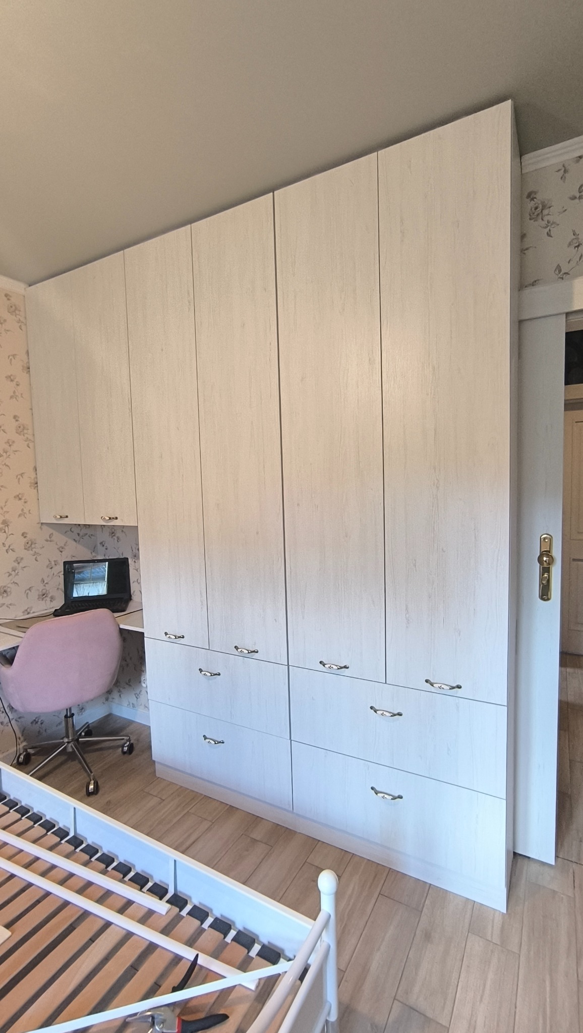 Jasna szafa wnękowa z szufladami i biurkiem w pokoju z tapetą w kwiaty. Widoczny fragment łóżka. Srebrne uchwyty. Ustawiona pod sufit.