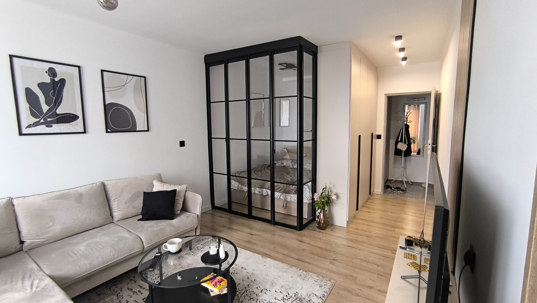 Nowoczesny salon z szklaną ścianką działową w czarnej ramie, oddzielającą sypialnię. Beżowa sofa, abstrakcyjne obrazy i drewniana podłoga tworzą spójną całość.