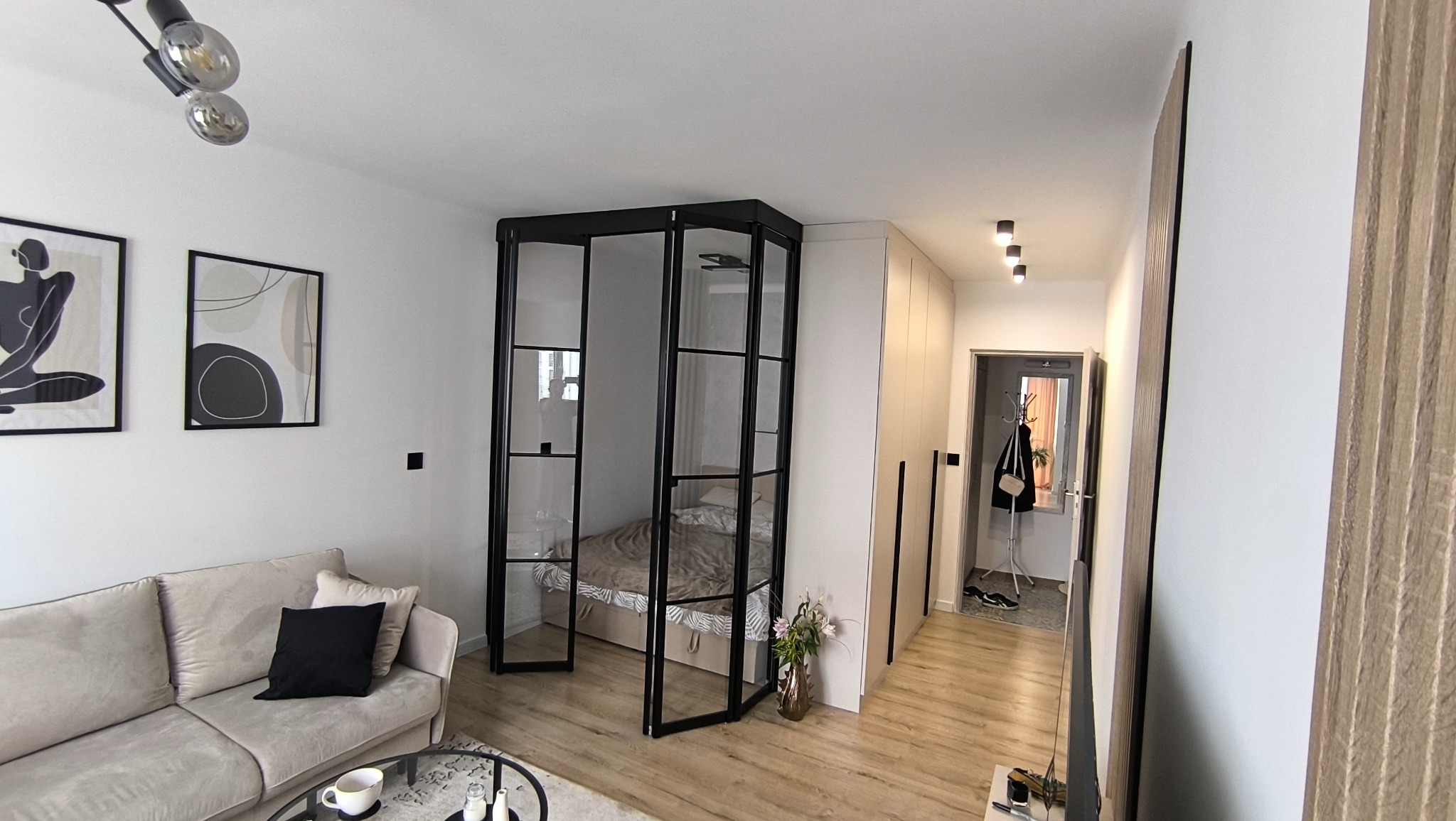 Nowoczesny pokój z wydzieloną strefą sypialnianą za pomocą szklanych drzwi w czarnej ramie. Minimalistyczny design, jasne kolory, drewniana podłoga i szafy w zabudowie.