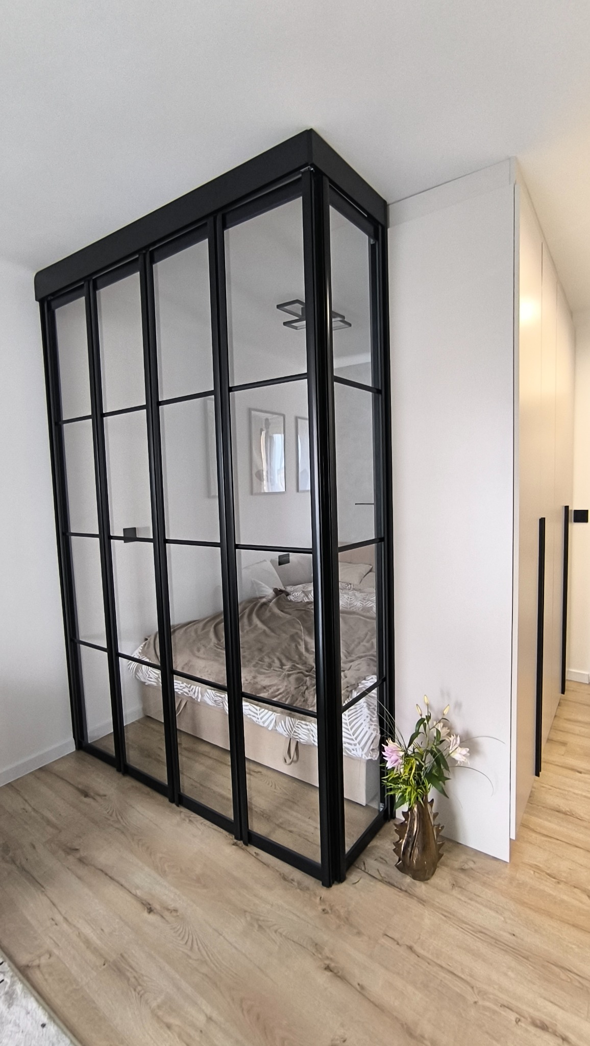 Loftowa, szklana ścianka działowa z czarnymi szprosami oddziela sypialnię od reszty mieszkania. Na podłodze panele, w rogu wazon z kwiatami. Minimalistyczny design.